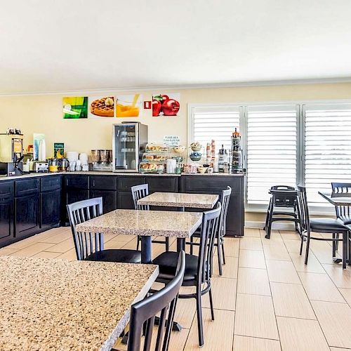 An indoor dining area with several tables and chairs, a beverage and snack station, and a counter with breakfast items and appliances.