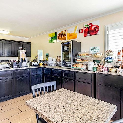 A small kitchen or pantry with dark cabinets, a table, coffee machines, and various food items and appliances on the counters.