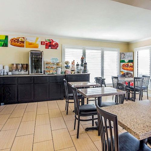 This image shows a bright dining area with several tables and chairs, a counter with food and beverage items, and large windows providing ample light.