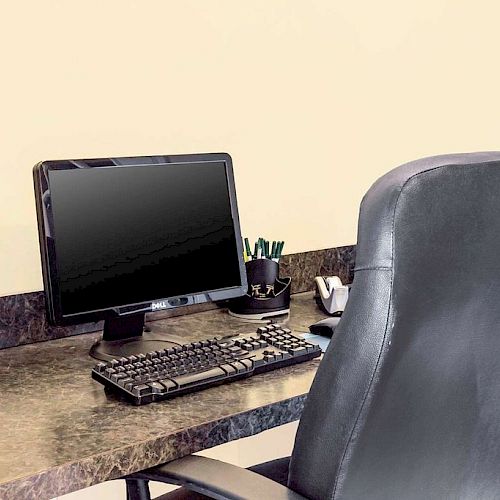 An office workspace with a computer, keyboard, and a black leather chair. There is a pen holder with pens next to the computer on the desk.