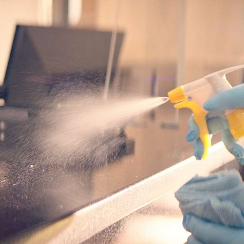 A person wearing gloves is disinfecting a surface with a spray bottle, while another person in the background is working at a computer.