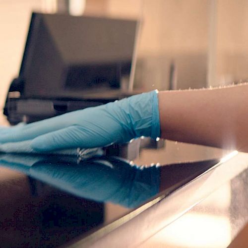 A close-up of a person cleaning a surface while wearing blue gloves, with a blurred figure in the background and a computer on a desk.