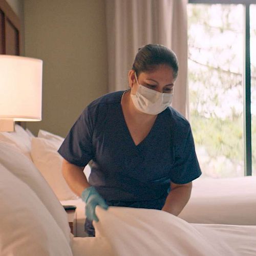 A person wearing a mask and gloves is making a bed in a hotel room with a lamp and window in the background.