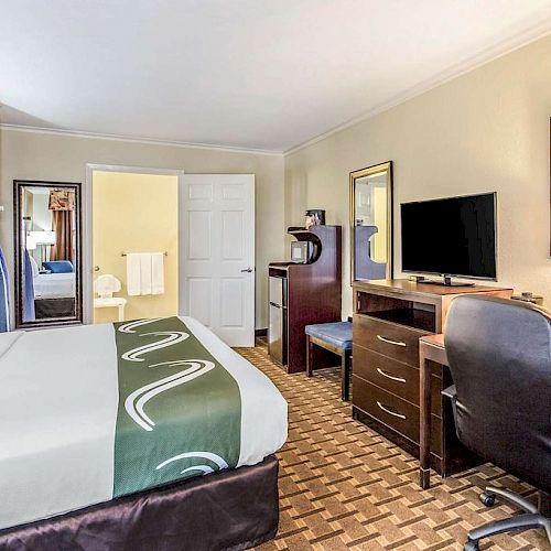 This image shows a hotel room with a bed, desk, chair, flat-screen TV, mirror, and a painting of horses. The door to the bathroom is open.