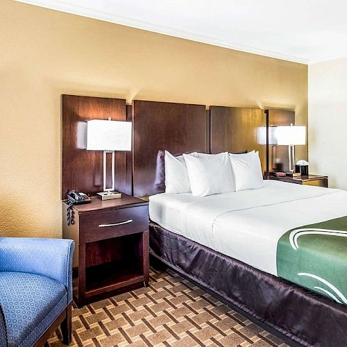 This image shows a hotel room with a bed, side tables with lamps, a blue armchair, phone, and an open closet with an iron and ironing board.