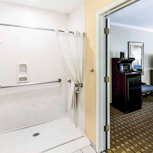 The image shows a bathroom with a walk-in shower with a grab bar, next to an open door leading to a room with furniture and a TV.