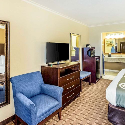 The image shows a hotel room with a bed, blue chair, TV, dresser, large mirror, and an open area with a microwave and coffee maker.