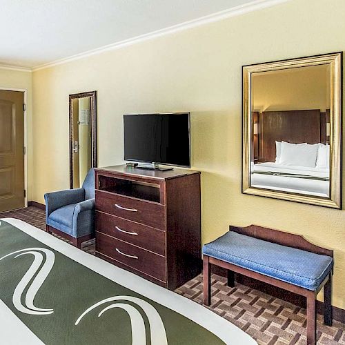 A hotel room features a bed, TV, chair, mirror, microwave, and small fridge. Decor is simple with cream-colored walls and wooden furniture.