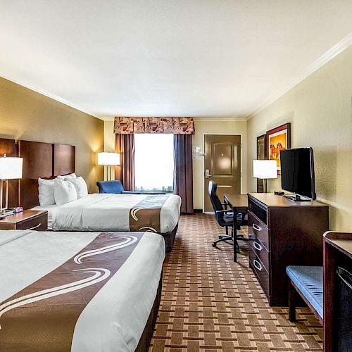 This image shows a hotel room with two double beds, a desk with a chair, a TV on a dresser, a mini-fridge, and a microwave.