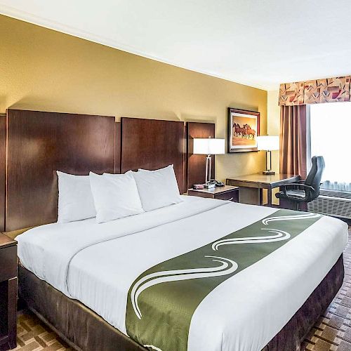 A hotel room with a king-sized bed, bedside tables with lamps, a desk and chair by the window, and neutral-colored decor.