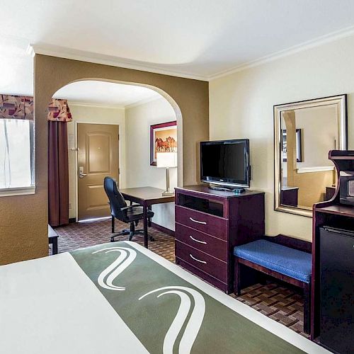 The image shows a hotel room with a bed, TV on a dresser, a mini-fridge, microwave, desk, chair, mirror, and an archway leading to a door.