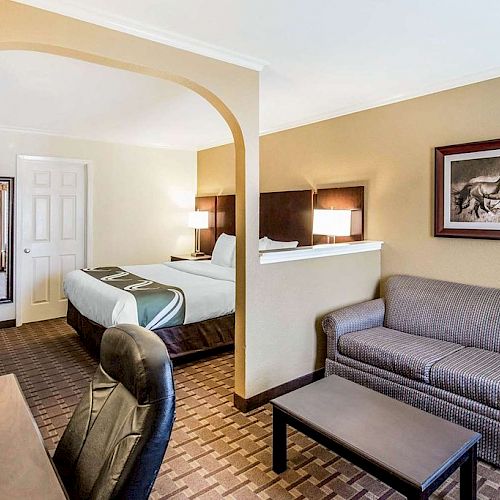 A hotel room features a bed, couch, desk, chair, and framed artwork. There's an archway partition, and a full-length mirror near the entrance door.