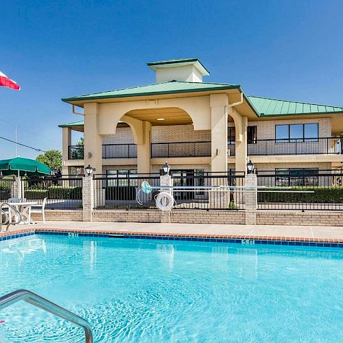 An outdoor swimming pool in front of a building with Texas and American flags, surrounded by patio furniture and a fence.