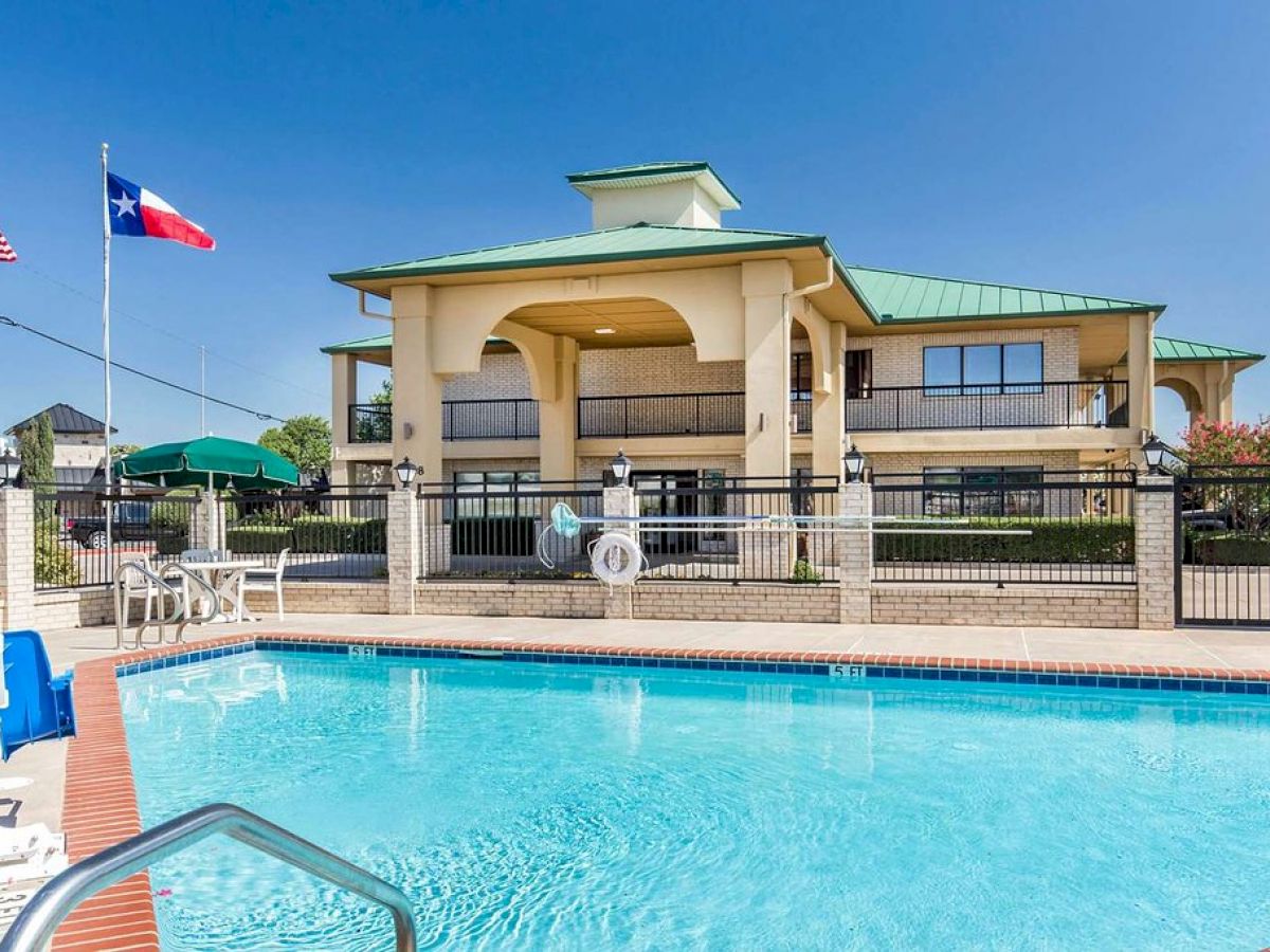 The image shows an outdoor swimming pool in front of a two-story building with a green roof, plus the U.S. and Texas flags.