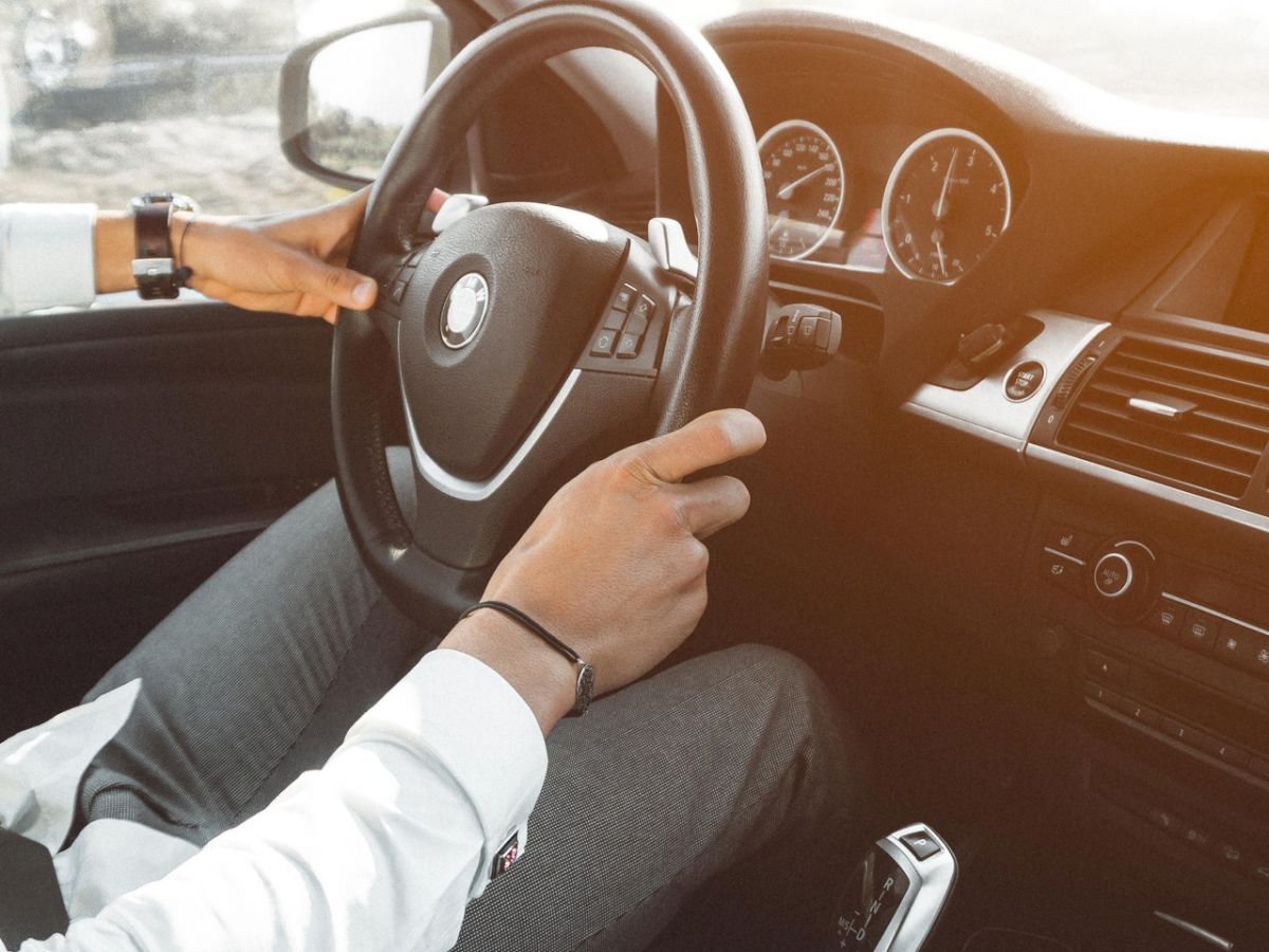 A person is driving a car, holding the steering wheel with both hands. The car interior is modern, and the driver's forearm is wearing a wristband.