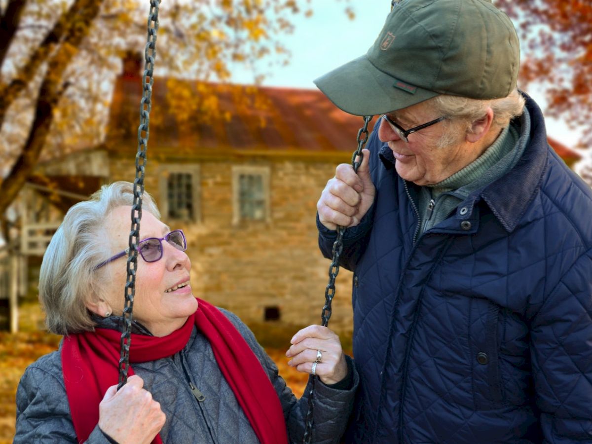 An elderly couple is enjoying time outdoors in an autumn setting with one of them sitting on a swing. They both seem happy and engaged.