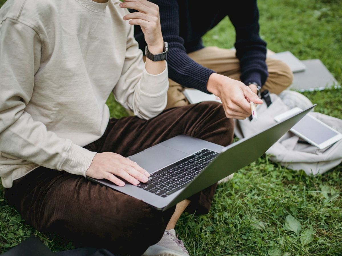 Two individuals are sitting on grass with a laptop and a tablet, engaged in discussion outdoors, one pointing at the screen.