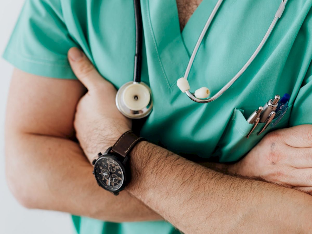 A person wearing green scrubs with a stethoscope around their neck, a wristwatch, and their arms crossed, possibly a medical professional.