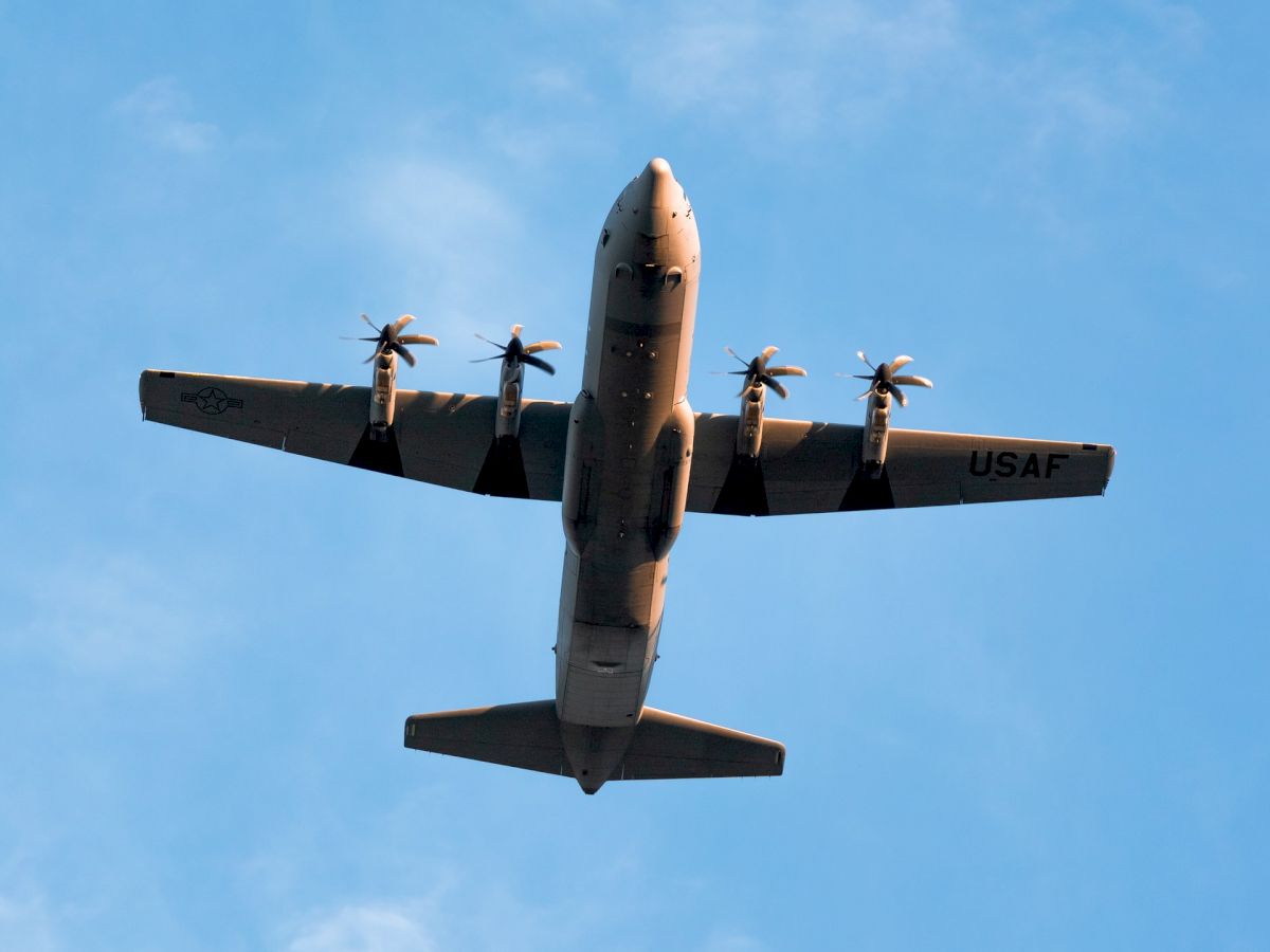 An image of a four-engine airplane flying overhead against a blue sky with scattered clouds.