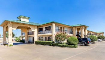 The image shows a two-story motel with a green roof, a parking lot, and several cars parked near the entrance, under a clear blue sky.