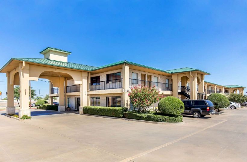 The image shows a two-story motel with a green roof, a parking lot, and several cars parked near the entrance, under a clear blue sky.