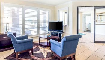 A cozy room with two blue armchairs, a TV on a stand, large windows with blinds, and a star-patterned rug. The room has an inviting ambiance.