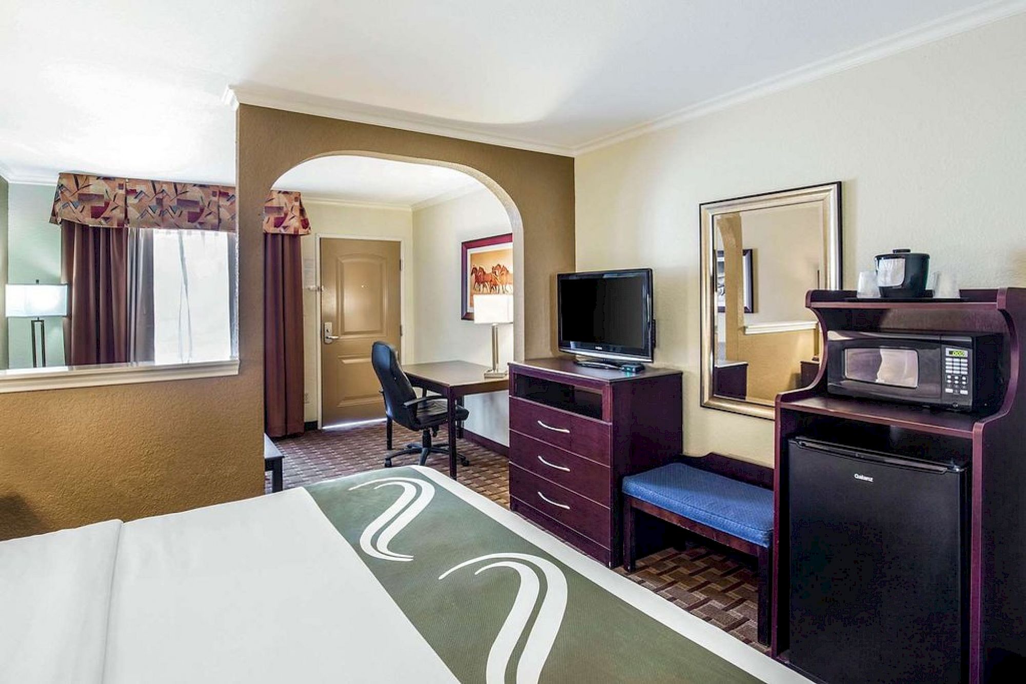 A hotel room with a bed, TV, dresser, desk, microwave, mini-fridge, and mirror, featuring an archway leading to another area with a door and window.