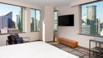 The image shows a modern hotel room with large windows offering a cityscape view, a flat-screen TV, a bed, and minimalist furnishings.