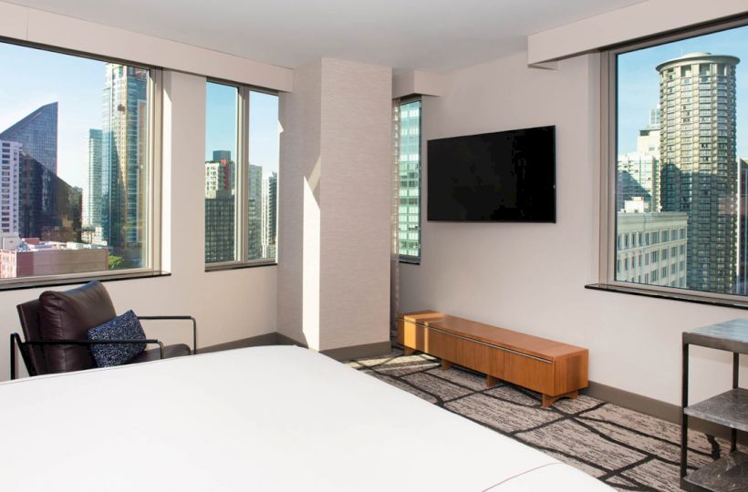 The image shows a modern hotel room with large windows offering a cityscape view, a flat-screen TV, a bed, and minimalist furnishings.