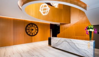 A modern lobby area with a curved wooden staircase, a large wall clock, a marble reception desk, and a bouquet of flowers ending the sentence.