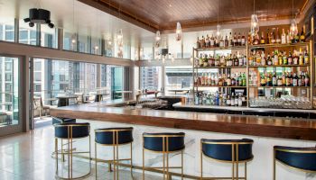 A modern bar interior featuring a long counter with black and gold stools, a well-stocked back bar, and large windows with a city view.