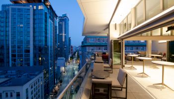 A modern cityscape at twilight, featuring tall buildings and a spacious balcony with seating and tables overlooking a lit urban street.