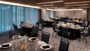 An elegantly set dining hall with round tables, black chairs, wine glasses, and folded napkins, under modern lighting, ready for an event.