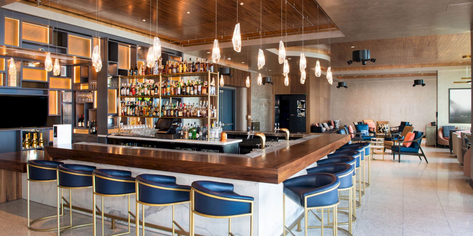 A modern bar with a wooden U-shaped counter, blue bar stools, hanging lights, and various bottles on shelves behind the counter.