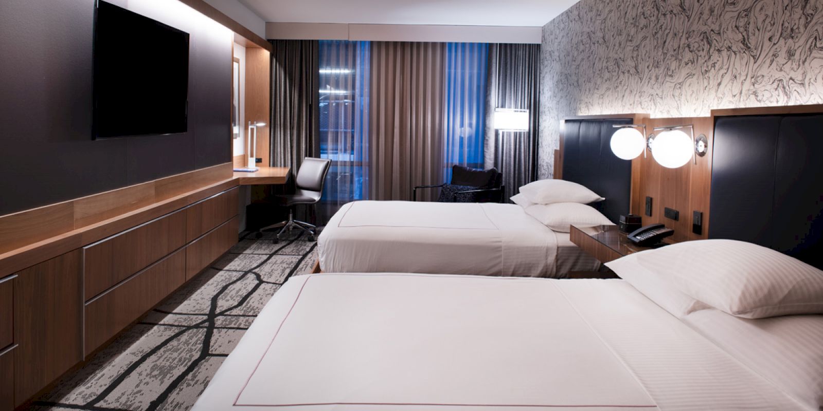 A modern hotel room featuring two neatly made beds, a wall-mounted TV, a desk with a chair, and patterned walls and carpeting.