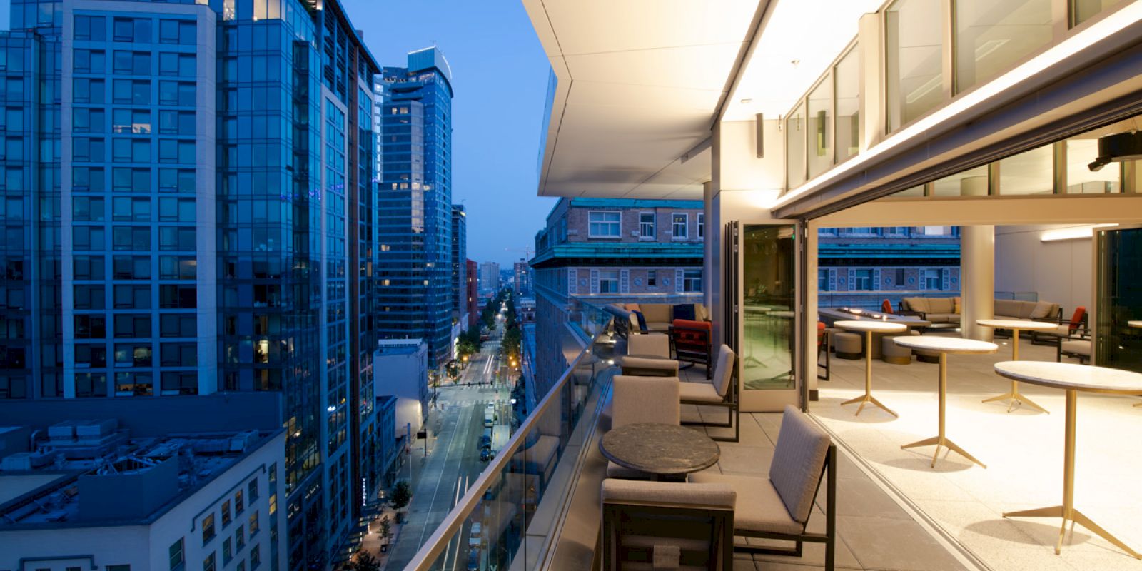 This image shows a rooftop terrace with tables and chairs, overlooking a cityscape with modern high-rise buildings during the evening.