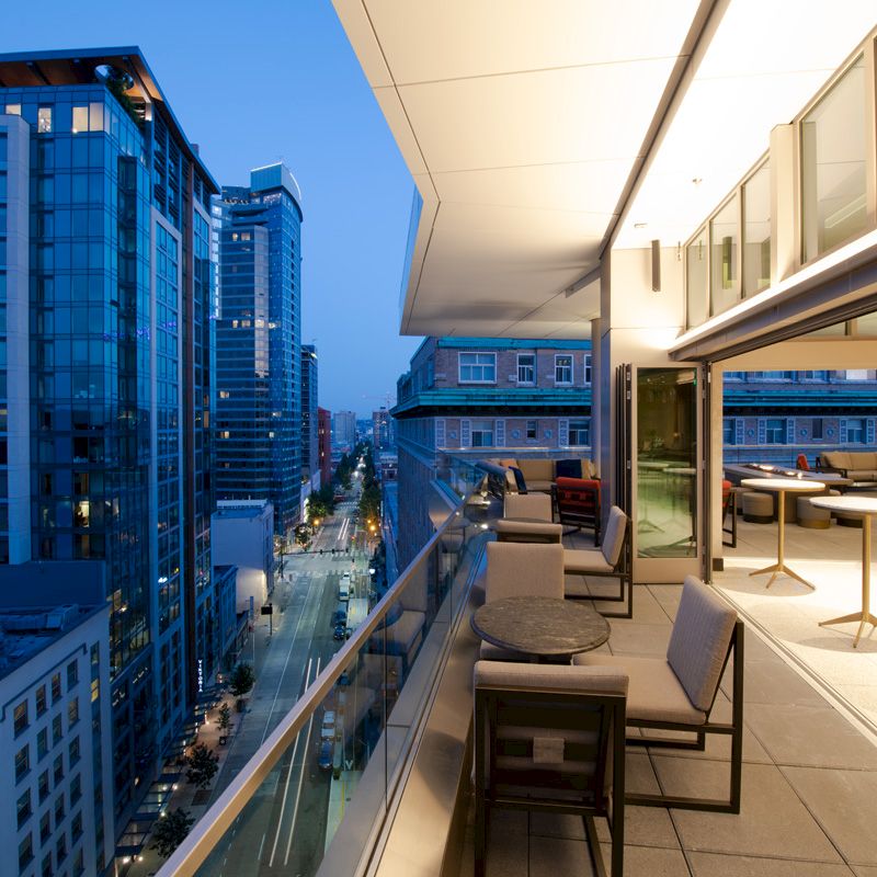 The image shows a modern rooftop terrace with seating areas, overlooking a cityscape with tall buildings and a busy street below, captured at dusk.