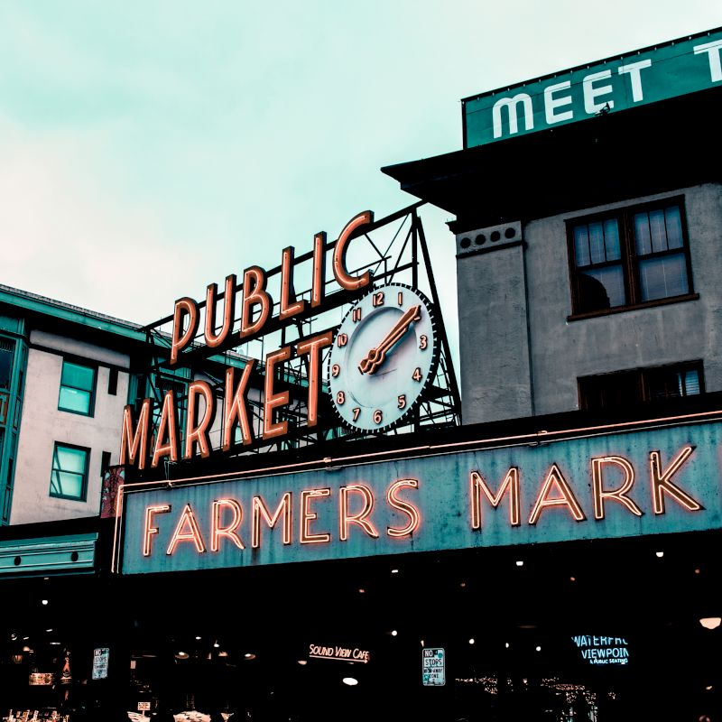 The image shows an outdoor scene with a "Public Market" sign featuring a clock and a "Farmers Market" sign below it on a vintage building.