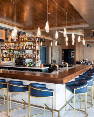 This image shows a modern bar with a wooden countertop, hanging lights, blue bar stools, and shelves stocked with various bottles.