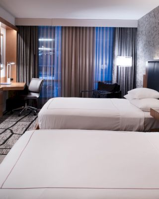 A modern hotel room features two neatly made beds, a flat-screen TV, a work desk in front of a window with curtains.