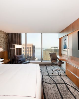A modern hotel room features a large bed, a wall-mounted TV, a desk with a chair, a patterned rug, and a view of buildings and water through large windows.