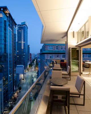 A modern cityscape at dusk, viewed from a sophisticated terrace with seating and tables, adjacent to a tall, glass-walled building.
