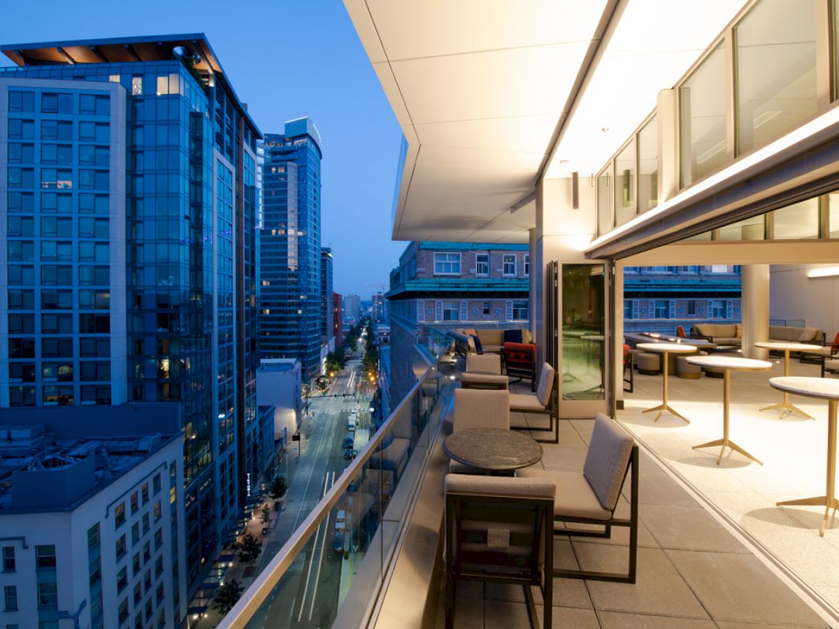 A modern balcony view, overlooking a city street lined with tall buildings, featuring tables and chairs, lit up in the early evening.