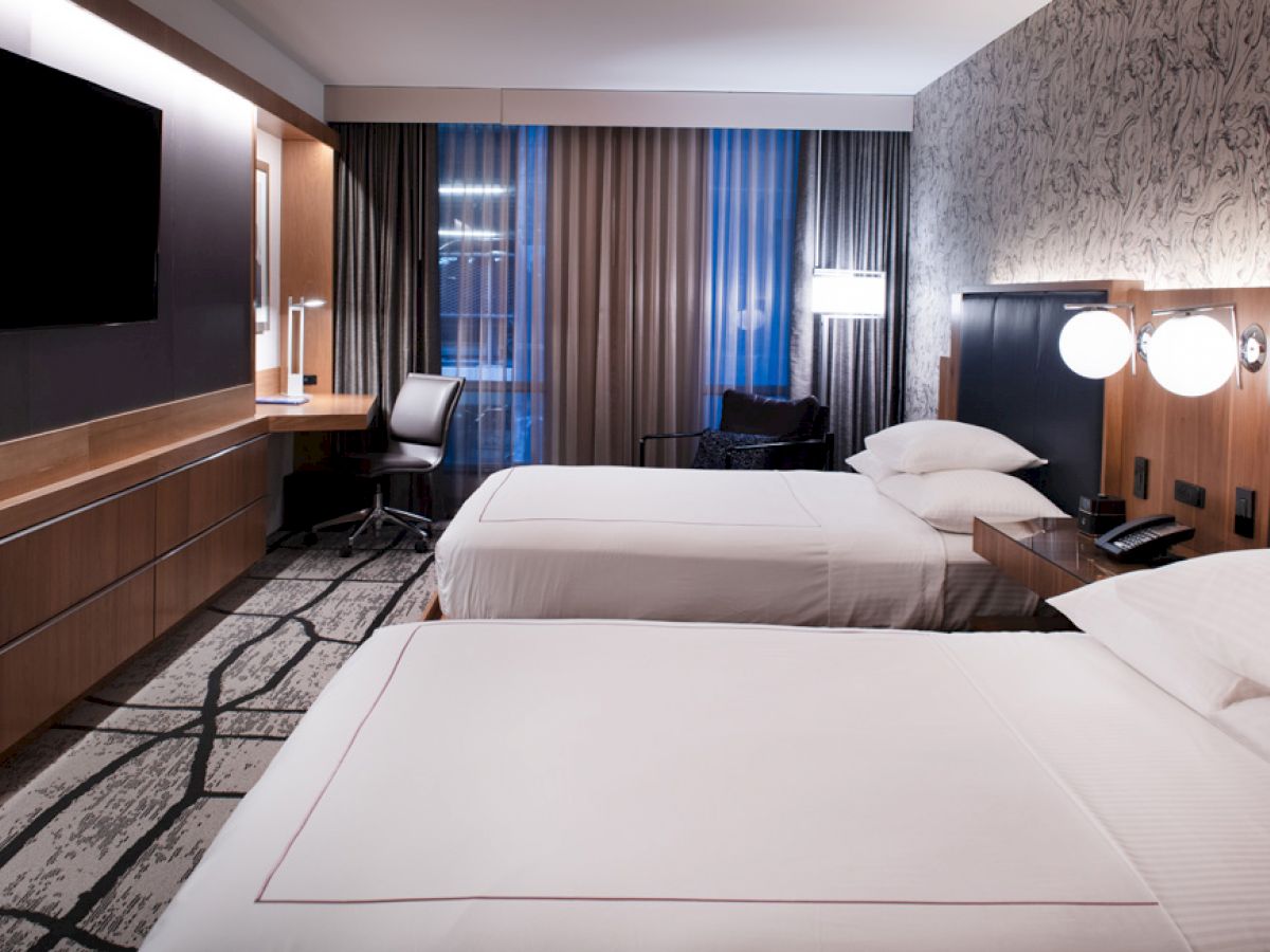 A modern hotel room with two neatly made beds, a wall-mounted TV, desk, chair, and large windows with curtains, providing a sleek and comfortable ambiance.