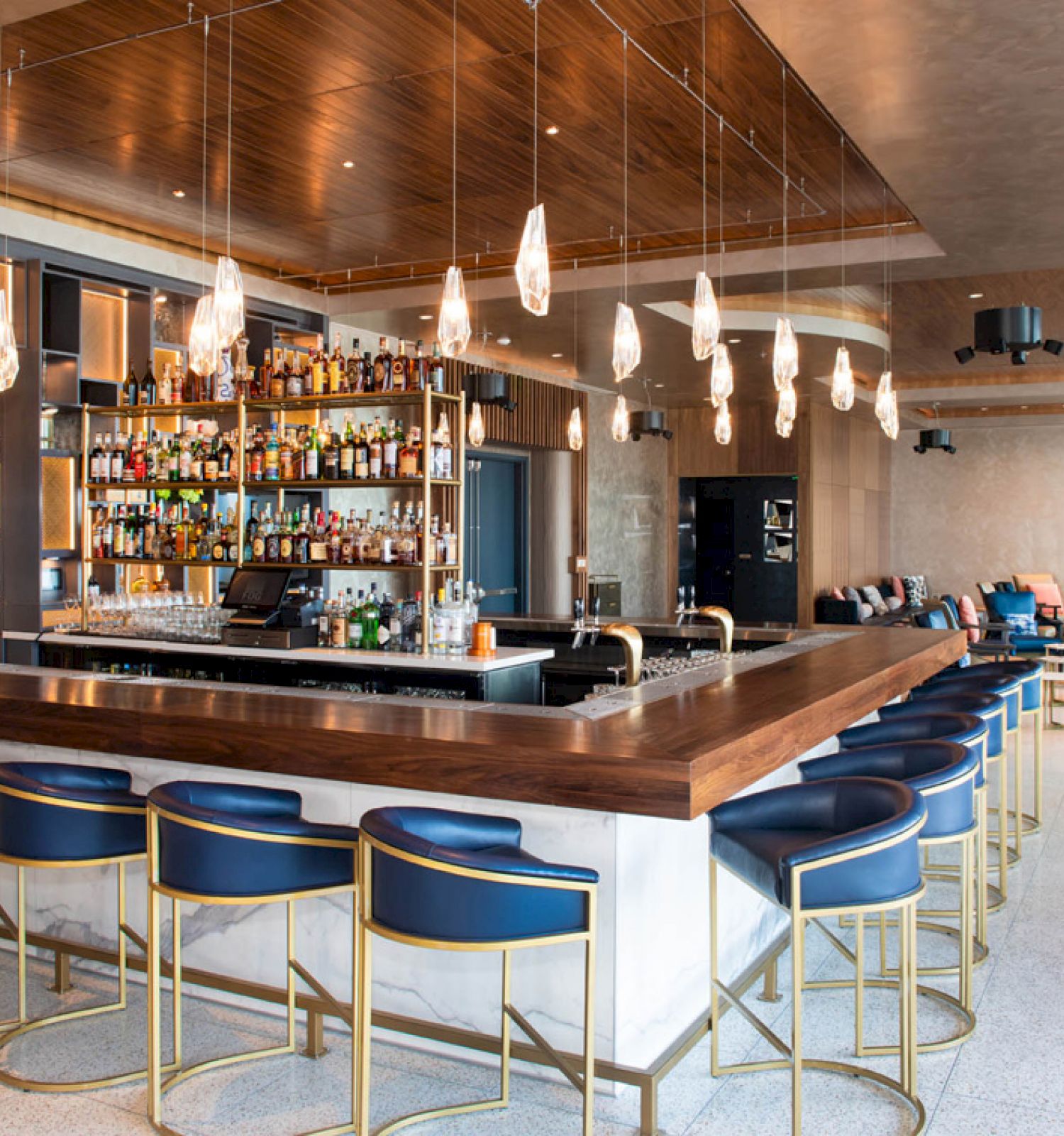 This image shows a modern bar with a well-stocked shelf, stylish seating, and hanging lights, surrounded by comfortable lounge areas.