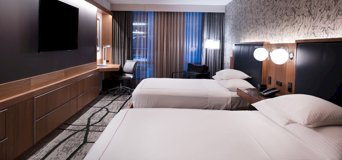 A modern hotel room features two neatly made beds, a work desk, a flat-screen TV, and stylish lighting.
