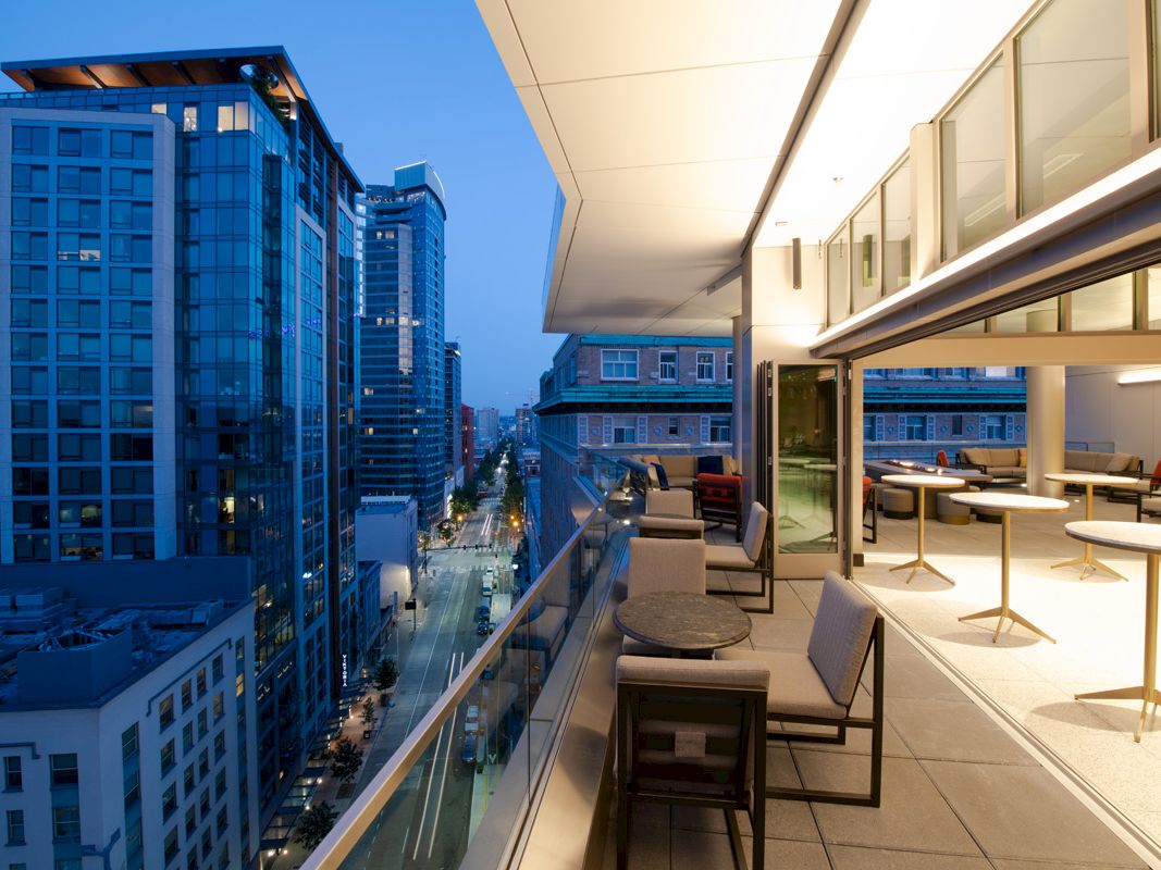 The image shows a modern cityscape with tall buildings and a stylish outdoor balcony area featuring seating and tables, all illuminated at dusk.