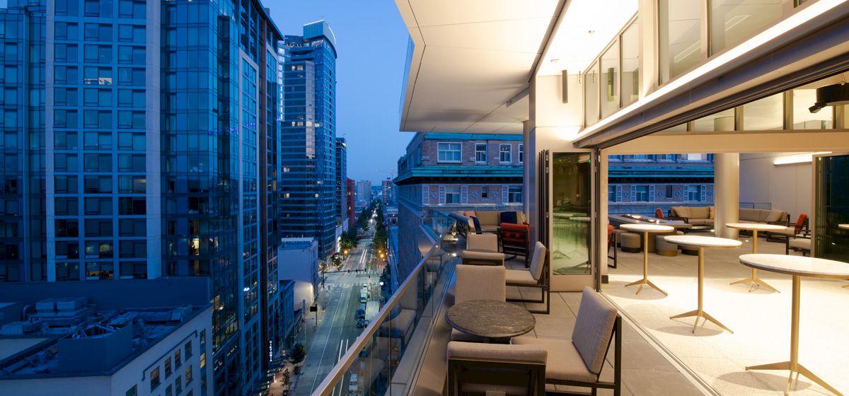 The image shows a modern cityscape with tall buildings and a stylish outdoor balcony area featuring seating and tables, all illuminated at dusk.