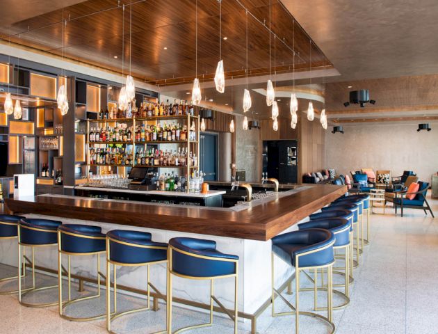 A modern bar with blue barstools, a well-stocked bar, and hanging lights. The room features contemporary decor and seating areas.