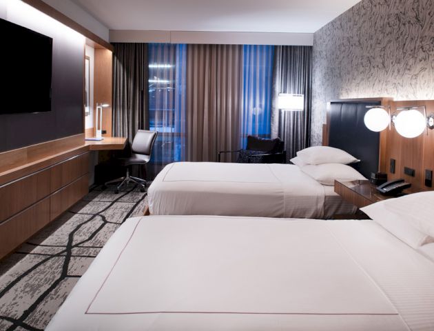 This image shows a modern hotel room with two beds, a TV, a desk, and a large window with curtains. The lighting is warm and inviting.
